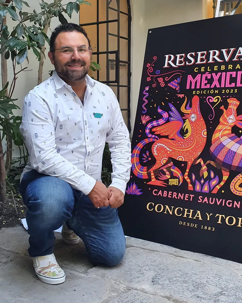 Mario Flores en cuclillas junto a un cartel grande de su etiqueta para Reservado Celebra México.