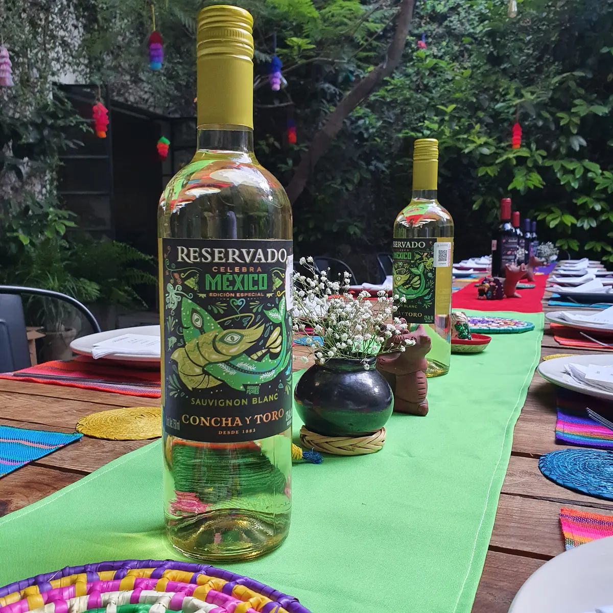 Botella de Sauvignon Blanc Reservado Celebra México con su etiqueta del pez verde, sobre una mesa de fiesta con camino verde y platos tejidos.
