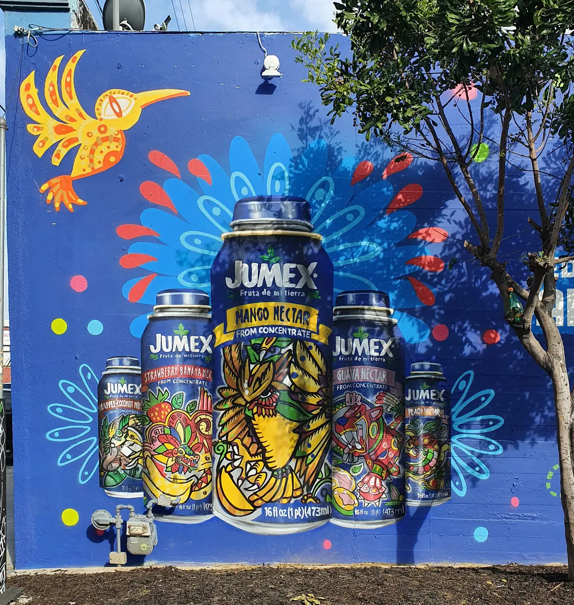 Mural de Jumex en Los Ángeles: latas gigantes de néctar de mango rodeadas de un colibrí y ornamentos sobre fondo azul.