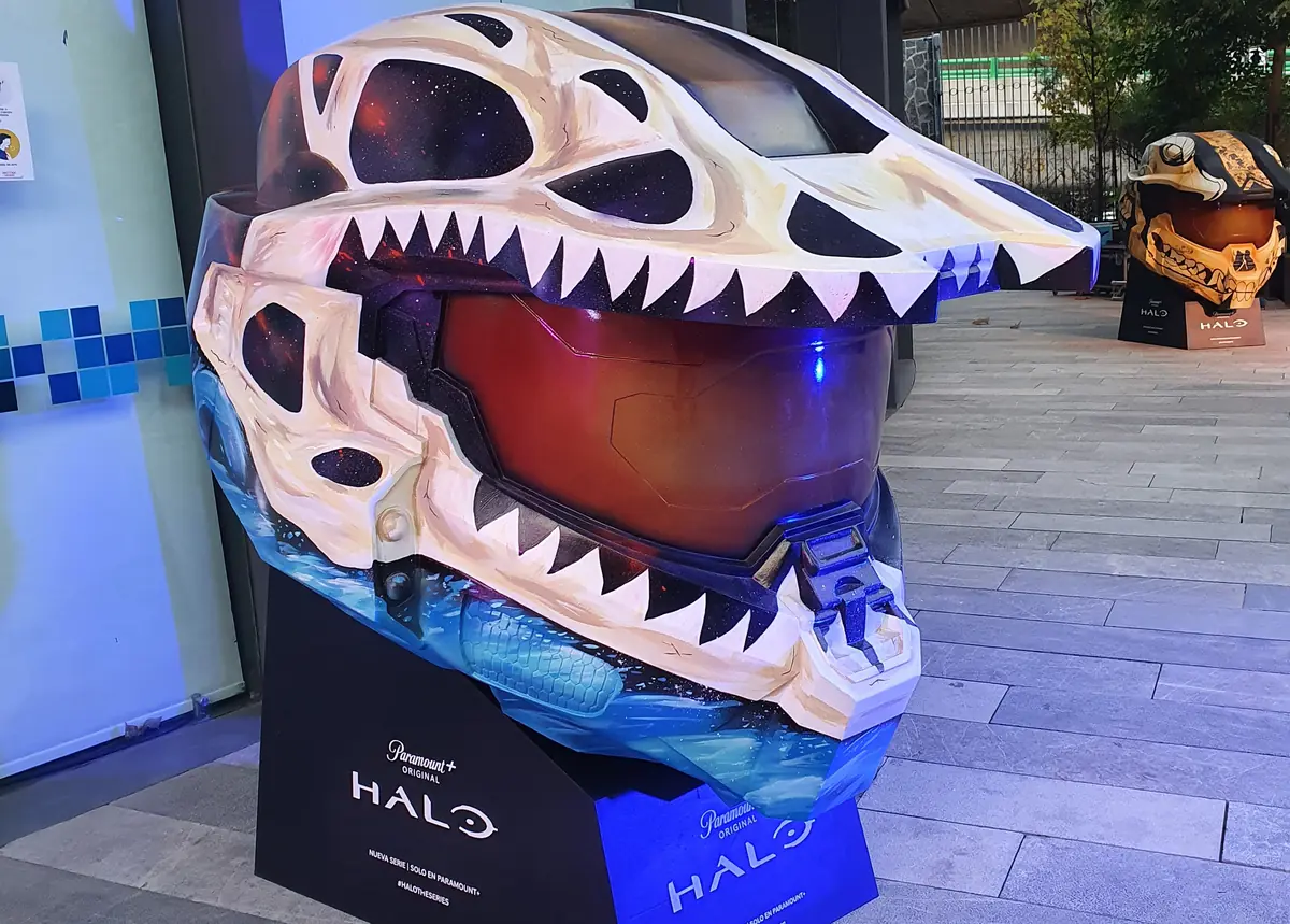 El casco exhibido de día en una plaza, sobre su base con el logo de Halo; al fondo, otro casco de la misma exposición.