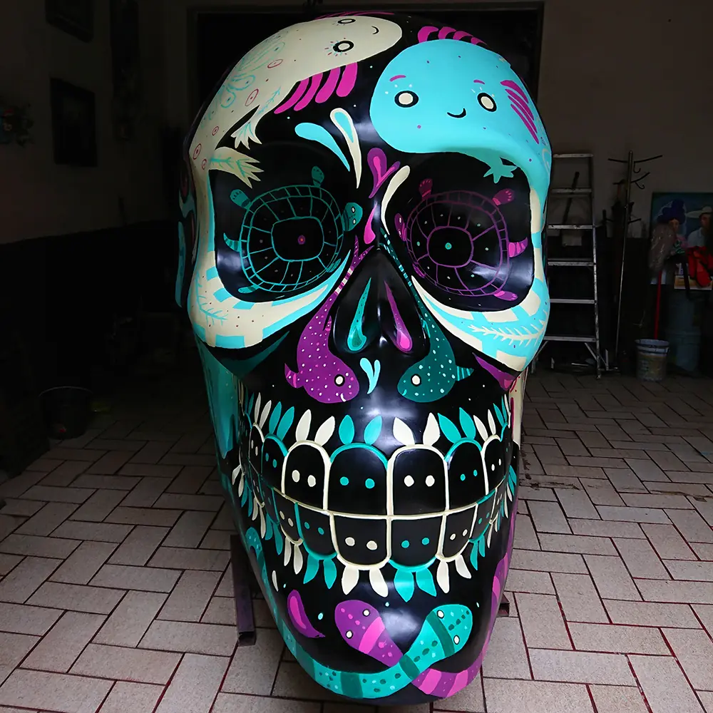Calavera monumental pintada de negro con ajolotes y criaturas en turquesa, rosa y blanco, vista de frente.